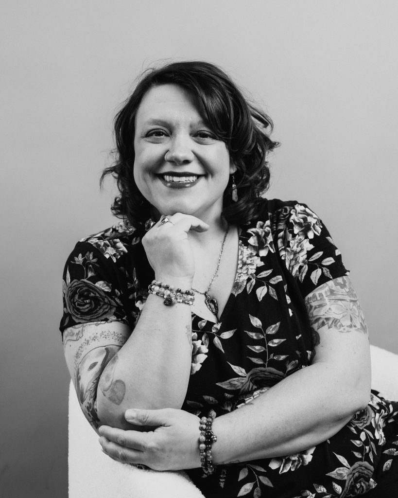 A black and white photo of Christina wearing a floral top, sitting in a white chair, smiling.